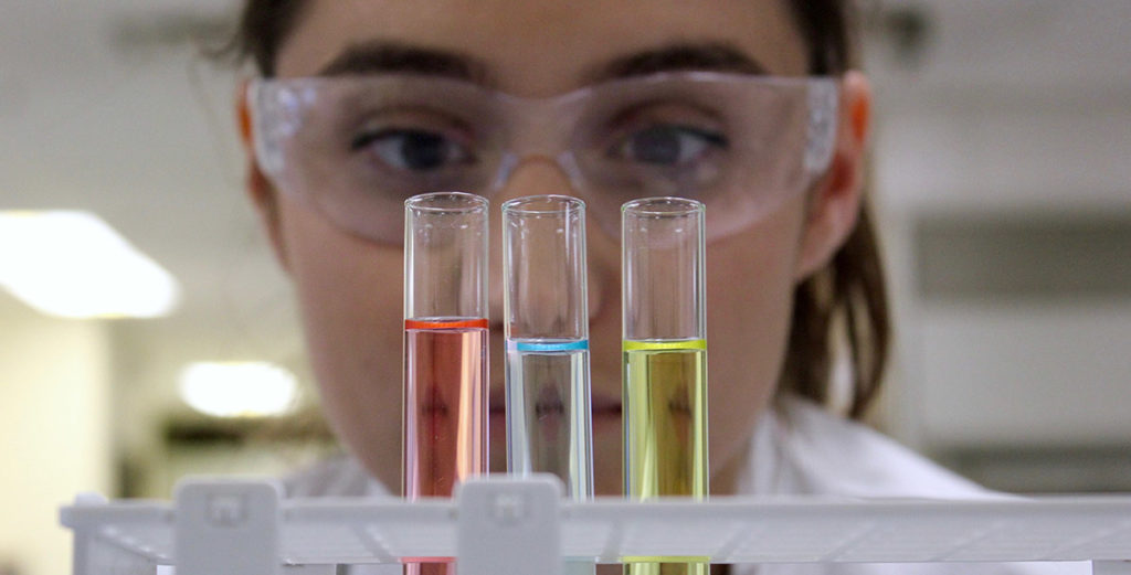 an image of a female lab worker in protective eyewear looking closely at 3 vials of different colored chemicals